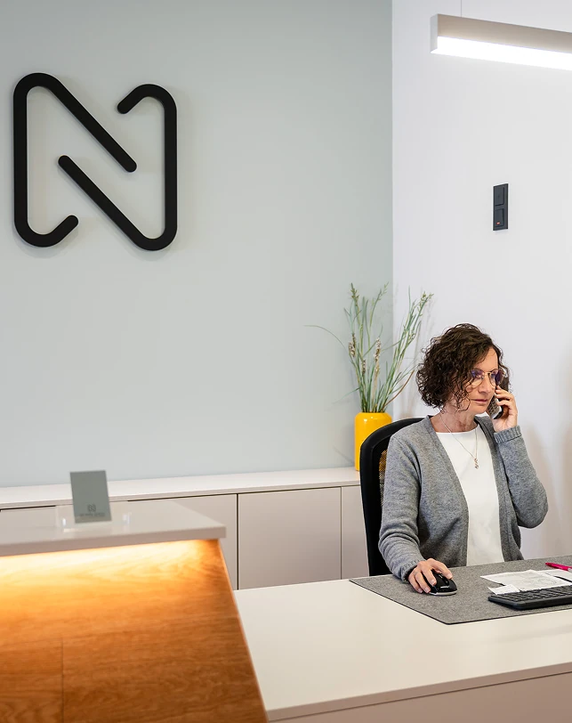 Frau mit lockigem Haar und Brille in grauer Strickjacke telefoniert an einem modernen Empfangstresen mit schwarzem Logo an der Wand sowie gelber Vase mit Pflanzen im Hintergrund.